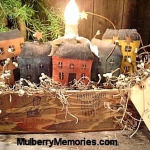 May include: A rustic wooden box holds several fabric houses in shades of orange, blue, and yellow, topped with a lit candle. The houses have small windows and doors. Twigs and white berries surround the houses, with a tag that reads "Home Sweet Home."