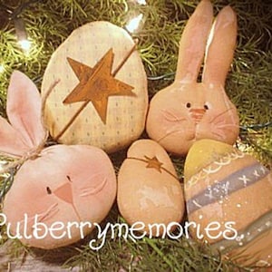 May include: A collection of Easter-themed decorations, including bunny and egg shapes, in soft pastel colors. The eggs feature star and stripe designs. The text "Mulberrymemories" is visible. The decorations are arranged on a bed of greenery with string lights.