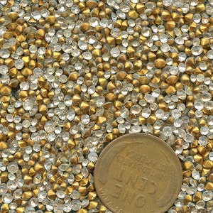 May include: A close-up shot of numerous gold and clear rhinestones. The rhinestones are small and scattered, with a penny for scale. The gems have a faceted appearance, reflecting light and creating a sparkling effect.