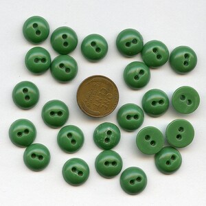 May include: A collection of twenty-one green, two-hole buttons scattered on a white surface. A copper-colored one-cent coin is also present, providing a size reference. The buttons are a solid, rounded design.