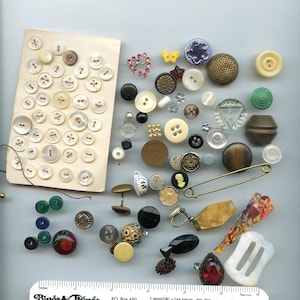 May include: A collection of assorted sewing buttons and fasteners, including a card of white buttons, a gold safety pin, a red rhinestone pendant, a yellow butterfly button, and various metal, plastic, and glass pieces, shown with a ruler.