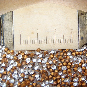 May include: Close-up of a ruler with measurements in centimetres, next to a pile of clear and copper-coloured rhinestones. The ruler is made of a light-coloured material and is marked with black lines.