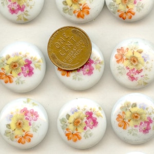 Könnte beinhalten: Eine Sammlung weißer, runder Keramik-Schrankknöpfe mit floralen Designs in Gelb, Orange und Rosa. Eine Kupfermünze liegt auf einem Knopf und zeigt die Worte "One Cent".