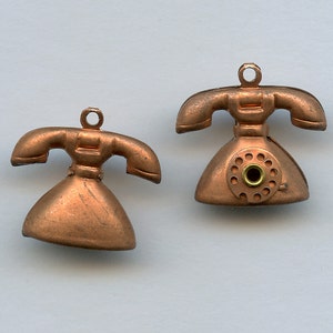 Pode incluir: Dois pingentes de telefone vintage cor de cobre. Cada pingente apresenta um mostrador rotativo e um auscultador. Os pingentes têm uma argola na parte superior para fixar em joias ou outros trabalhos manuais. Os pingentes de telefone são um design retrô.