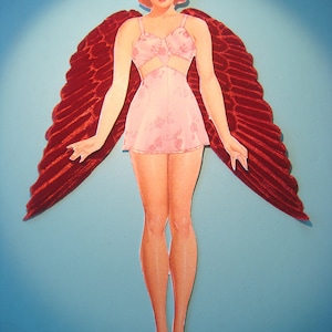 May include: A vintage paper doll cutout of a woman with red hair, wearing a pink floral dress and red velvet wings. The doll is posed against a light blue background.