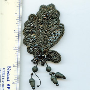 May include: A black lace applique featuring intricate corded floral patterns and dark bead embellishments. The piece includes two dangling corded tassels with beads. A ruler placed alongside shows the applique measures approximately 4.5 inches long.