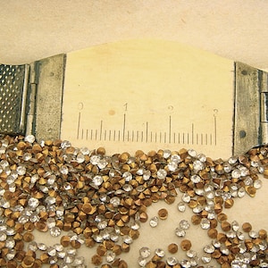 May include: A collection of small, faceted rhinestones in gold and clear colors. A vintage ruler with a metal edge is positioned above the rhinestones. The ruler shows measurements in inches.
