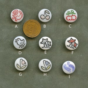 May include: A collection of nine white, round buttons with various hand-drawn designs, including a rooster, cherries, mushrooms, a bird, and flowers. A US penny is included for size reference. The buttons are labeled A through I.