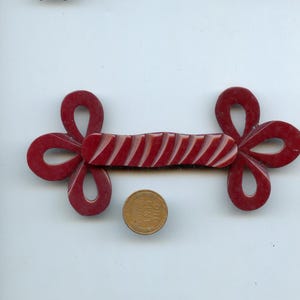 Può includere: Un fermaglio a levetta decorativo rosso scuro con una sezione centrale scolpita e scanalata e quattro estremità ad anello. Una moneta da un centesimo è posta sotto per il confronto delle dimensioni. La chiusura è probabilmente per un capo di abbigliamento o un accessorio.