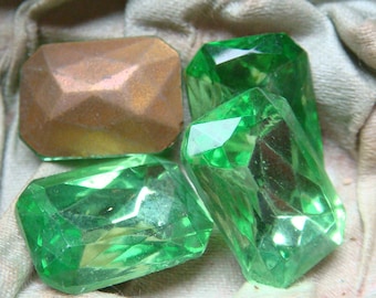 May include: Four faceted gemstones. Three are a vibrant green, and one is a bronze color. The gemstones have a rectangular shape with cut edges. The gems are displayed on a textured, light-colored fabric.