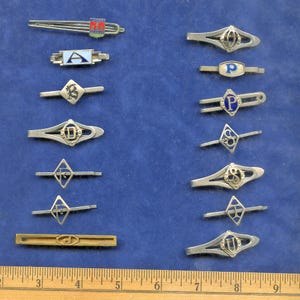 Peut inclure: Collection de pinces à cravate vintage argentées et dorées, avec diverses lettres initiales et motifs. Certaines pinces ont des détails en émail bleu, rouge et blanc. Les pinces sont disposées sur une surface bleue, avec une règle en bas.