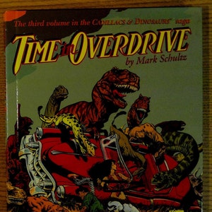 May include: A comic book cover featuring a red car with dinosaurs and humans. The title of the comic is "Time Overdrive" by Mark Schultz. The third volume in the "Cadillacs & Dinosaurs" saga.