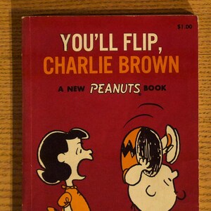May include: A vintage book cover for "You'll Flip, Charlie Brown," featuring Lucy and Charlie Brown from the Peanuts comic strip. The cover is a deep red with white and orange text, and illustrations of the characters.