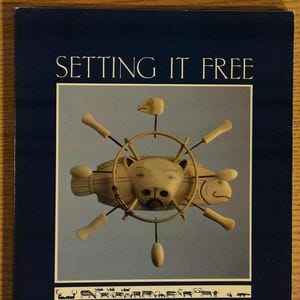 May include: A book cover with the title "SETTING IT FREE" in white text. The cover features a light-colored carving of a fish-like creature with a bear-like face, surrounded by a wheel-like structure. The bottom text reads "An Exhibition of Modern Alaskan Eskimo Ivory Carving."
