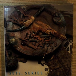 May include: A book cover with a brown background and the title "Sets, Series & Ensembles in African Art". The cover features a close-up of a wooden bowl filled with various African artifacts, including a wooden figure, a spear, and a woven mat.