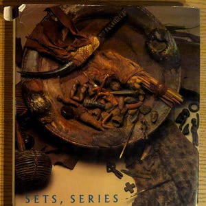 May include: A book cover titled "SETS, SERIES & ENSEMBLES IN AFRICAN ART." The cover features a detailed, textured image of various African artifacts, including a bowl, horn, and small sculptures, in shades of brown and tan.