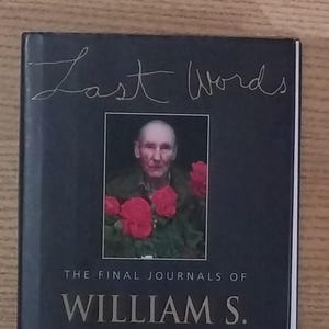 May include: A hardcover book titled "Last Words" with a portrait of a man surrounded by red flowers. The book is a collection of journals by William S. Burroughs, edited by James Grauerholz. The cover is dark gray.