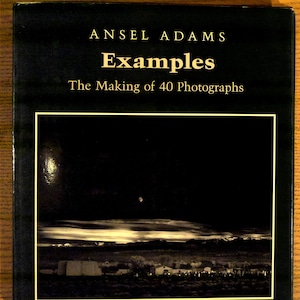 May include: A hardcover book titled "Examples" by Ansel Adams. The cover is black with gold lettering and a sepia-toned photograph of a landscape with a night sky and a small town. The book is about the making of 40 photographs.
