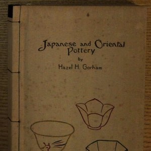 May include: The cover of a book titled "Japanese and Oriental Pottery" by Hazel H. Gorham. The cover features three illustrations of pottery: a red flower-shaped bowl, a white bowl with a black and red design, and a black octagonal bowl with a white design.