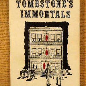 May include: Vintage book cover titled "Tombstone's Immortals." The cover features a black and white illustration of a three-story building with figures on balconies and in front. Red accents highlight the building's details. The title is in bold, black letters.