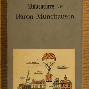 May include: A vintage book titled "The Surprising Adventures of Baron Munchausen." The cover is gray with the title in black text. A colorful illustration depicts a hot air balloon over a town.