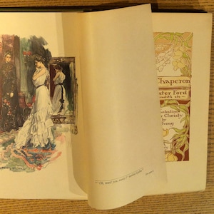 May include: Open book with illustrations. One page shows a woman in a white dress, standing in front of a mirror. The opposite page has the title "Chaperon" and the author's name. The book is bound in green.