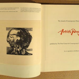 May include: Open book with a black and white print of a person's head on the left page. The right page has red text that reads "Artist's Proof" and "The Annual of Contemporary Prints, Volume VII."