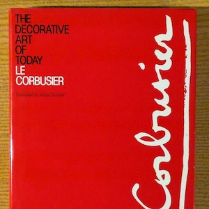 May include: A red book cover with the title "The Decorative Art of Today" and the author "Le Corbusier" in black text. The author's name is also written in white in a large, stylized signature on the right side of the cover.