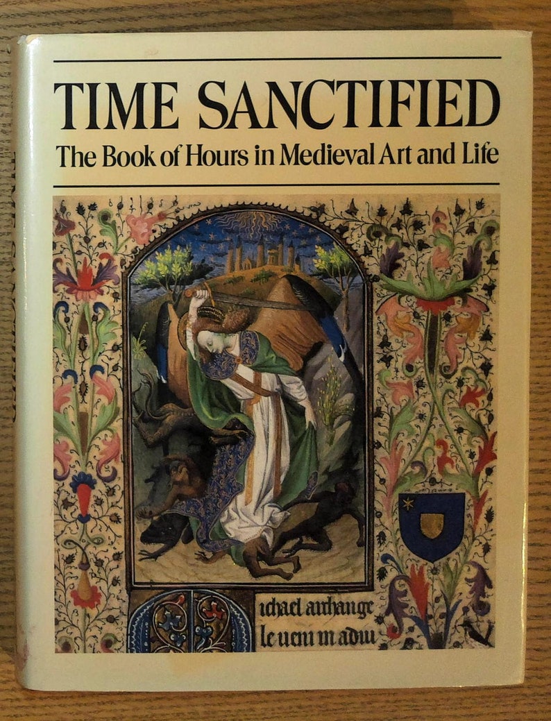 Puede incluir: Un libro de tapa dura titulado "TIME SANCTIFIED" con el subt&iacute;tulo "The Book of Hours in Medieval Art and Life". La portada presenta una ilustraci&oacute;n medieval con figuras, bordes florales y un escudo de armas. El libro est&aacute; sobre una superficie de madera.