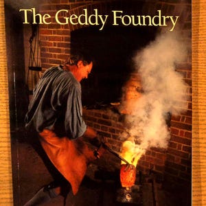 May include: A book cover titled The Geddy Foundry featuring a photograph of a person in a blue shirt and orange apron working with molten metal in a brick furnace, with bright orange flames and white smoke rising from the casting process.