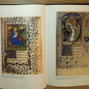 Puede incluir: Libro abierto con dos p&aacute;ginas de manuscritos iluminados. La p&aacute;gina izquierda presenta a la Virgen Mar&iacute;a y el ni&ntilde;o con una t&uacute;nica azul, rodeados de estrellas doradas y bordes florales. La p&aacute;gina derecha muestra una escena religiosa con figuras y dise&ntilde;os florales.