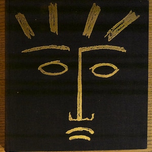 May include: A black book cover with a gold abstract face design. The face features oval eyes, a straight nose, and a mouth with two curved lines. Above the eyes are gold brushstroke-like shapes, suggesting a sun or radiating light.