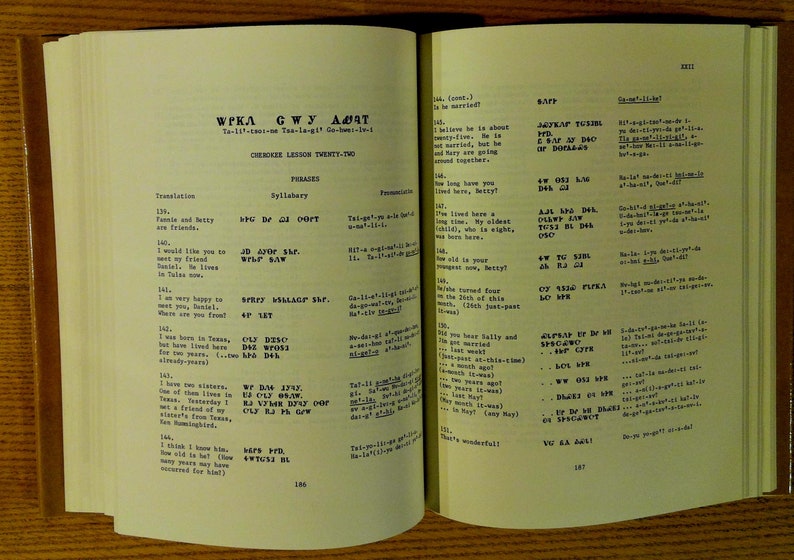 Może przedstawiać: Strona z książki zatytułowanej "WPKA GWY ADAT" z podtytułem "CHEROKEE LESSONS TWENTY-TWO" i tekstem "Ta-11-cao-nea-to-." Strona zawiera tłumaczenie fraz w języku czirokeeskim i odpowiadający im sylabariusz.