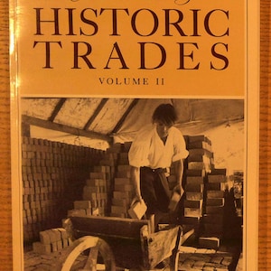 May include: A softcover book titled Colonial Williamsburg HISTORIC TRADES VOLUME II. The cover features a sepia-toned photograph of a person in period clothing loading bricks into a wooden wheelbarrow in a brickyard with stacks of bricks.