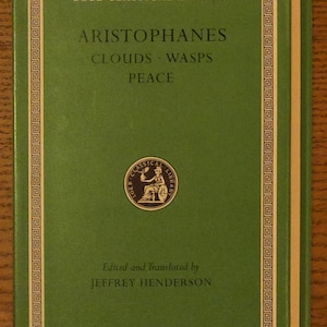 May include: A green hardcover book titled Loeb Classical Library Aristophanes Clouds Wasps Peace edited and translated by Jeffrey Henderson. The cover features a gold border and a circular emblem of a seated figure in the center with gold text.