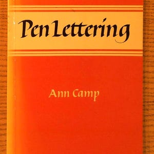 May include: A rectangular book with a bright orange cover featuring a white horizontal band across the top third. The text Pen Lettering is printed in black serif font within the band, and the author name Ann Camp is in white below.