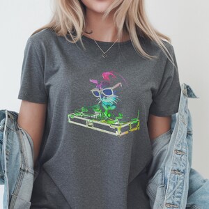 Op de afbeelding: Een donkergrijs t-shirt met een kleurrijke afbeelding van een kat-DJ met een zonnebril en een koptelefoon. De kat staat achter een DJ-mixer met draaitafels. Het ontwerp bevat levendige neonkleuren.