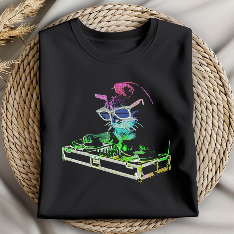 Op de afbeelding: Zwart T-shirt met een kleurrijke afbeelding van een kat met een zonnebril die draait op een DJ-draaitafel. De kat en de draaitafel zijn weergegeven in neonkleuren, waaronder groen, blauw en roze. Het T-shirt is opgevouwen op een geweven mat.