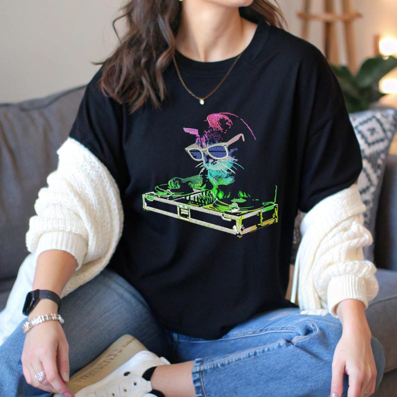 Op de afbeelding: Zwart t-shirt met een kleurrijke afbeelding van een kat met een zonnebril en koptelefoon, die draait op een DJ-tafel. De kat is weergegeven in tinten groen, blauw en roze. Het ontwerp is gecentreerd op de voorkant van het shirt.