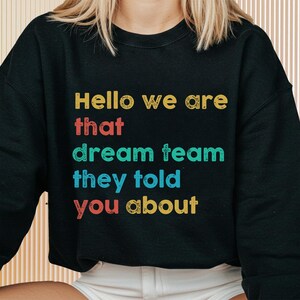 Puede incluir: Sudadera negra con el texto "Hello we are that dream team they told you about" en letras amarillas, rojas, turquesas y amarillas. La sudadera es de cuello redondo.