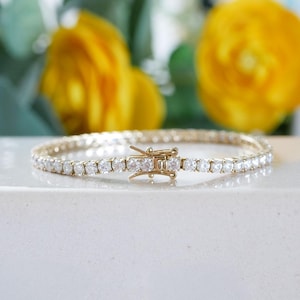 Pu&ograve; includere: Un braccialetto tennis color oro con numerose pietre trasparenti e scintillanti. Il braccialetto &egrave; chiuso e poggia su una superficie bianca, con fiori gialli e fogliame verde sullo sfondo.