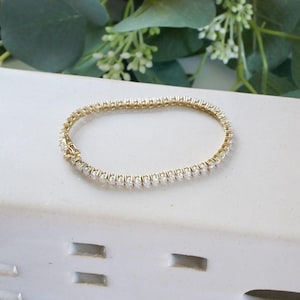 Pu&ograve; includere: Un braccialetto tennis dorato con una fila di pietre preziose chiare e scintillanti. Il braccialetto &egrave; esposto su una superficie bianca con vegetazione decorativa sullo sfondo. Un gioiello classico.
