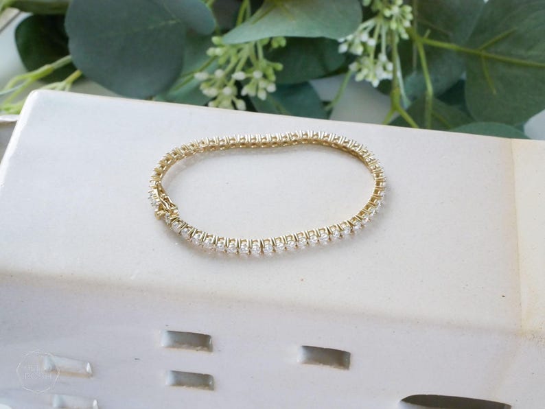 Pu&ograve; includere: Un braccialetto tennis dorato con una fila di pietre preziose chiare e scintillanti. Il braccialetto &egrave; esposto su una superficie bianca con vegetazione decorativa sullo sfondo. Un gioiello classico.