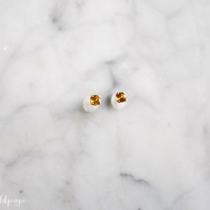 May include: A pair of small, gold stud earrings with a single, round, orange gemstone in each earring.