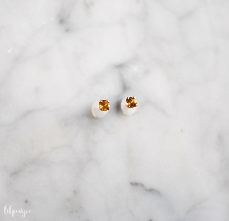 May include: A pair of small, gold stud earrings with a single, round, orange gemstone in each earring.