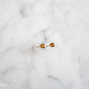 May include: A pair of gold stud earrings with small, oval-shaped, orange gemstones.