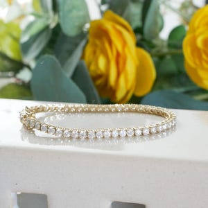 Pu&ograve; includere: Un bracciale dorato con una fila di pietre chiare e scintillanti. Il bracciale &egrave; appoggiato su una superficie bianca, con fiori gialli e fogliame verde sullo sfondo. Gioielli.