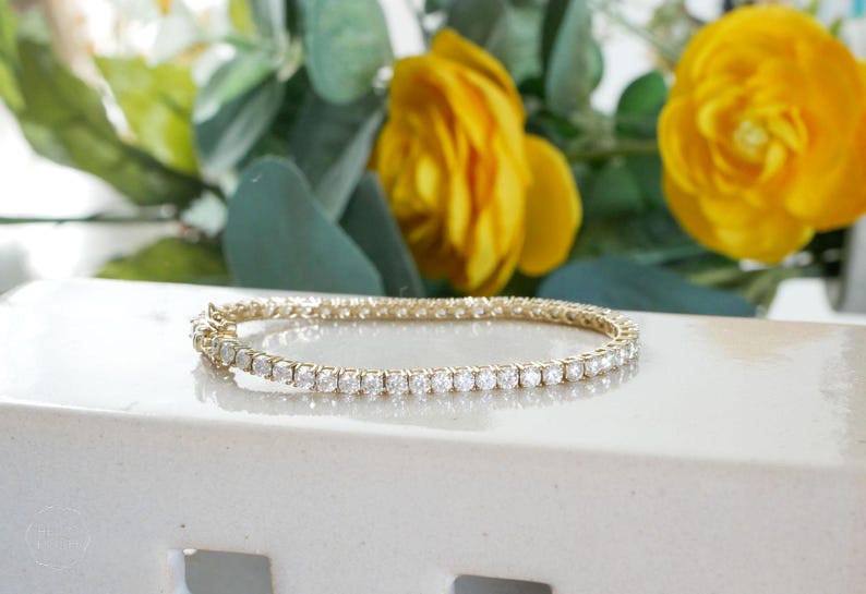 Pu&ograve; includere: Un bracciale dorato con una fila di pietre chiare e scintillanti. Il bracciale &egrave; appoggiato su una superficie bianca, con fiori gialli e fogliame verde sullo sfondo. Gioielli.