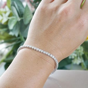 Pu&ograve; includere: Un braccialetto tennis color argento, con una fila continua di pietre preziose rotonde e trasparenti. Il braccialetto &egrave; indossato su un polso. Lo sfondo &egrave; sfocato, con verde e un tocco di fiori rosa.