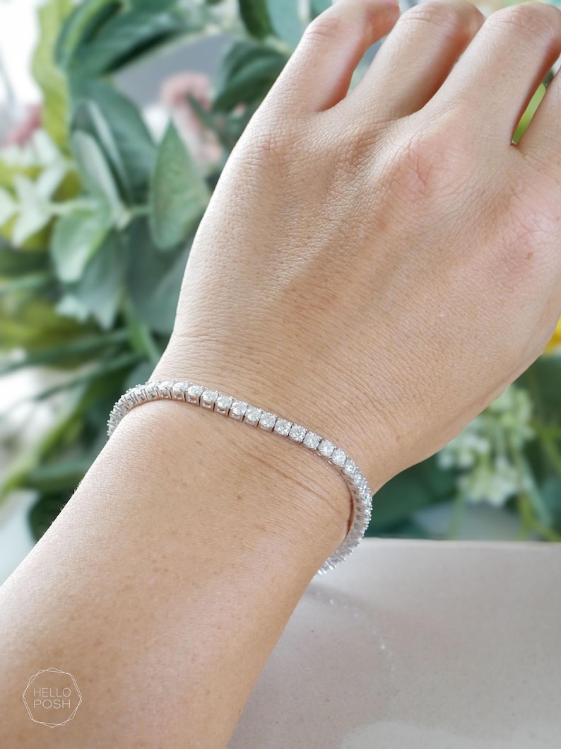 Pu&ograve; includere: Un braccialetto tennis color argento, con una fila continua di pietre preziose rotonde e trasparenti. Il braccialetto &egrave; indossato su un polso. Lo sfondo &egrave; sfocato, con verde e un tocco di fiori rosa.
